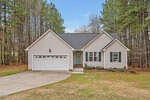 A single-story house with beige siding, dark shutters, a two-car garage, and a concrete driveway, set in front of a wooded area with pine trees under a blue sky.