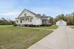 A white single-story house with a front porch and screened side porch sits on a spacious lot with a driveway leading to a detached white garage. The lawn is mostly grass, bordered by trees in the background.