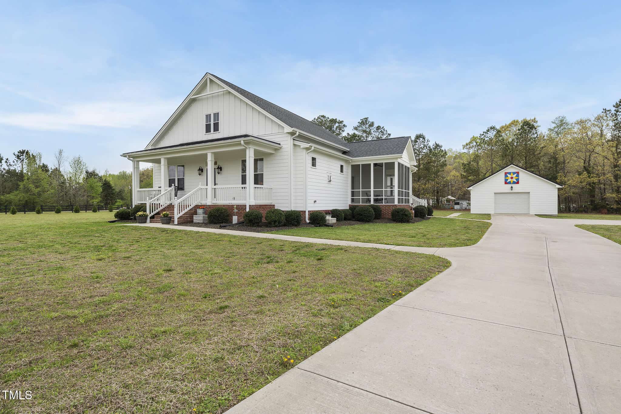 A white single-story house with a front porch and screened side porch sits on a spacious lot with a driveway leading to a detached white garage. The lawn is mostly grass, bordered by trees in the background.