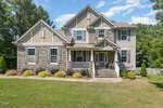 A two-story suburban house with stone and gray siding, white trim, a covered front porch, and landscaped yard with bushes and grass, surrounded by trees under a blue sky.