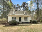 A small white house with a front porch, two dormer windows on the roof, green shutters, and bushes along the porch. The house is surrounded by trees and sits on a patchy lawn under a partly cloudy sky.