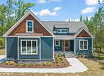 A modern single-story house with blue siding, brown shingles, and white trim, featuring large windows, a stone foundation, front steps, and a landscaped yard, surrounded by trees under a partly cloudy sky.