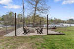 Four cushioned chairs are arranged around a fire pit on a stone patio by a lake, enclosed by a low fence. A large tree stands nearby, with docks and boats visible in the background under a blue sky.
