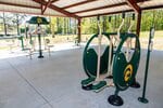 Outdoor covered area with green and beige exercise equipment on a concrete floor. The equipment includes elliptical machines and other fitness stations, surrounded by trees and grassy areas.