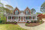 A two-story brick house with white trim, black shutters, and a covered front porch with columns. Potted plants and ferns decorate the porch. A curved driveway leads to the front steps. Trees and shrubs surround the home.