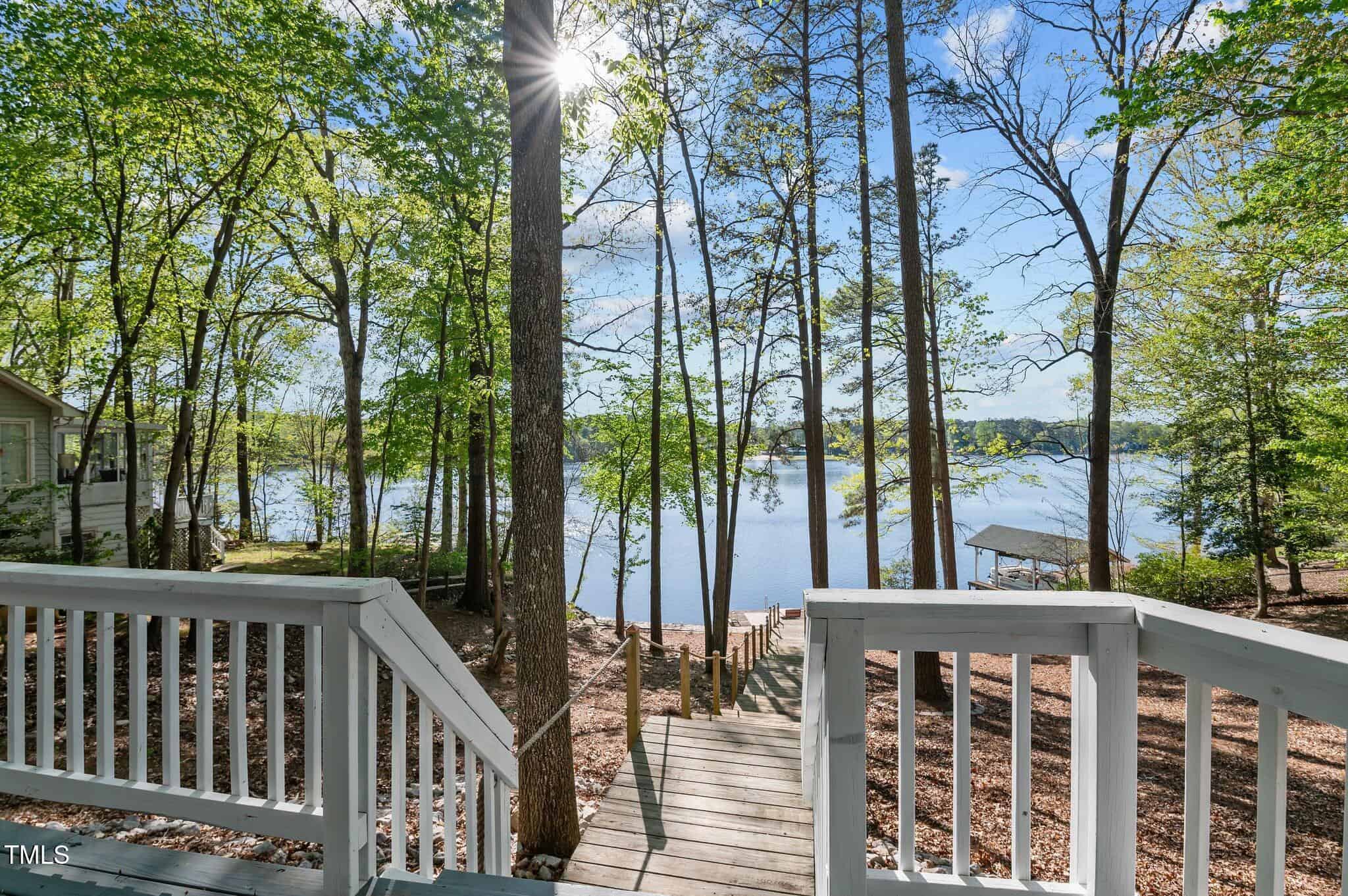 View from a white wooden deck with stairs leading down to a dock on a calm lake, surrounded by tall trees under a clear sky with sunlight streaming through the branches.