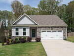 A single-story house with light-colored siding and stone accents, two front windows, a dark front door, a double garage, and a sloped driveway. The yard has green grass, bushes, and trees in the background.