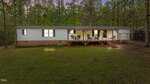 A single-story white manufactured home with green shutters and a brick foundation, featuring a covered wooden porch with outdoor furniture, set in a grassy yard surrounded by tall trees.