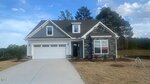 A two-story house with gray siding and stone accents, a double garage, and a "Sold" sign in the front window. The house sits on a grassy lot with small trees and a clear sky in the background.
