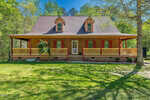A wooden house with a large covered front porch and dormer windows sits on a grassy lawn, surrounded by trees and greenery under a clear sky.