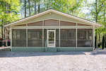 A single-story house with light green siding and a screened-in front porch, set on a gravel lot and surrounded by tall trees in a wooded area.
