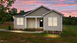 A single-story beige house with white trim and a covered front porch is shown at sunset. The house has a black front door, brick foundation, and sits on a grassy lot with trees in the background.
