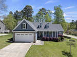 A light blue suburban house with white trim, two dormer windows, and a two-car garage. The front yard has a manicured lawn, bushes, and a concrete driveway. Tall trees and neighboring homes are visible in the background.