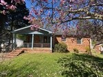 A single-story brick house with a small covered porch, surrounded by green grass and a flowering tree with pink blossoms in the foreground. The day is sunny with clear blue skies.
