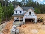A two-story house under construction with exterior walls wrapped in building paper, an unfinished front porch, construction materials stacked nearby, and an excavator parked next to a dirt lot surrounded by trees.
