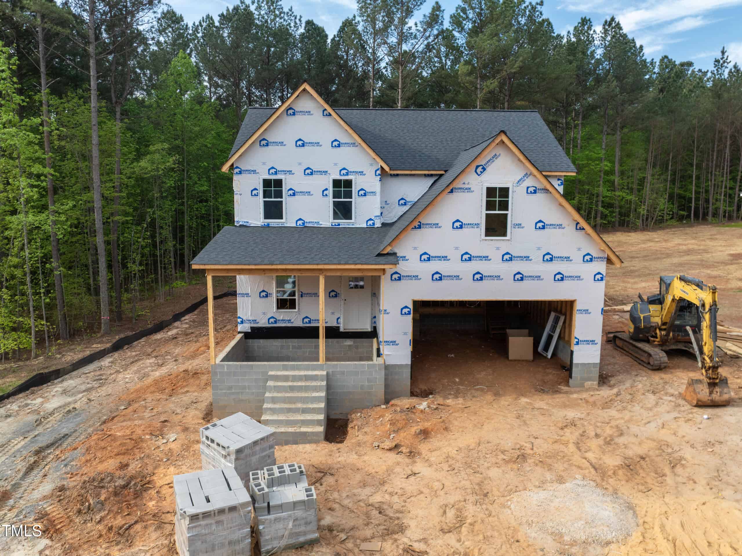 A two-story house under construction with exterior walls wrapped in building paper, an unfinished front porch, construction materials stacked nearby, and an excavator parked next to a dirt lot surrounded by trees.