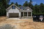 A newly constructed two-story house with a front porch, stone and siding exterior, and an attached garage. The driveway is unpaved, and a large dumpster sits to the right of the house. Trees are in the background.