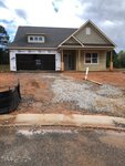 A newly constructed house with beige siding, a covered porch, and a double garage, with a "SOLD" sign in the window. The yard is unfinished with gravel and exposed dirt.
