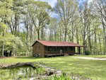 A one-story brick house with a red roof sits in a grassy clearing surrounded by tall trees. A small pond is in the foreground, and a gravel driveway leads up to the house. The sky is partly cloudy.