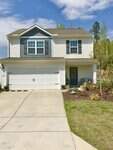 Two-story suburban house with white siding, gray shutters, and a double garage. A concrete driveway leads to the garage, and there are landscaped plants and a welcome wreath on the teal front door. Blue sky with clouds above.