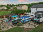 A backyard with a swimming pool, hot tub, and covered bar area with colorful stools. Several lounge chairs, string lights, a patio table, and landscaped garden beds surround the pool. Nearby houses and trees are visible.