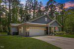 A single-story gray house with a double garage, American and another flag hanging by the entrance, set among tall trees and greenery at dusk. A concrete driveway leads up to the garage.