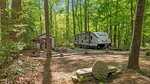 A white Bullet travel trailer is parked in a wooded campsite with a small shed, a covered swing, and a round stone sun sculpture in the foreground. Tall trees surround the area, and sunlight filters through the leaves.