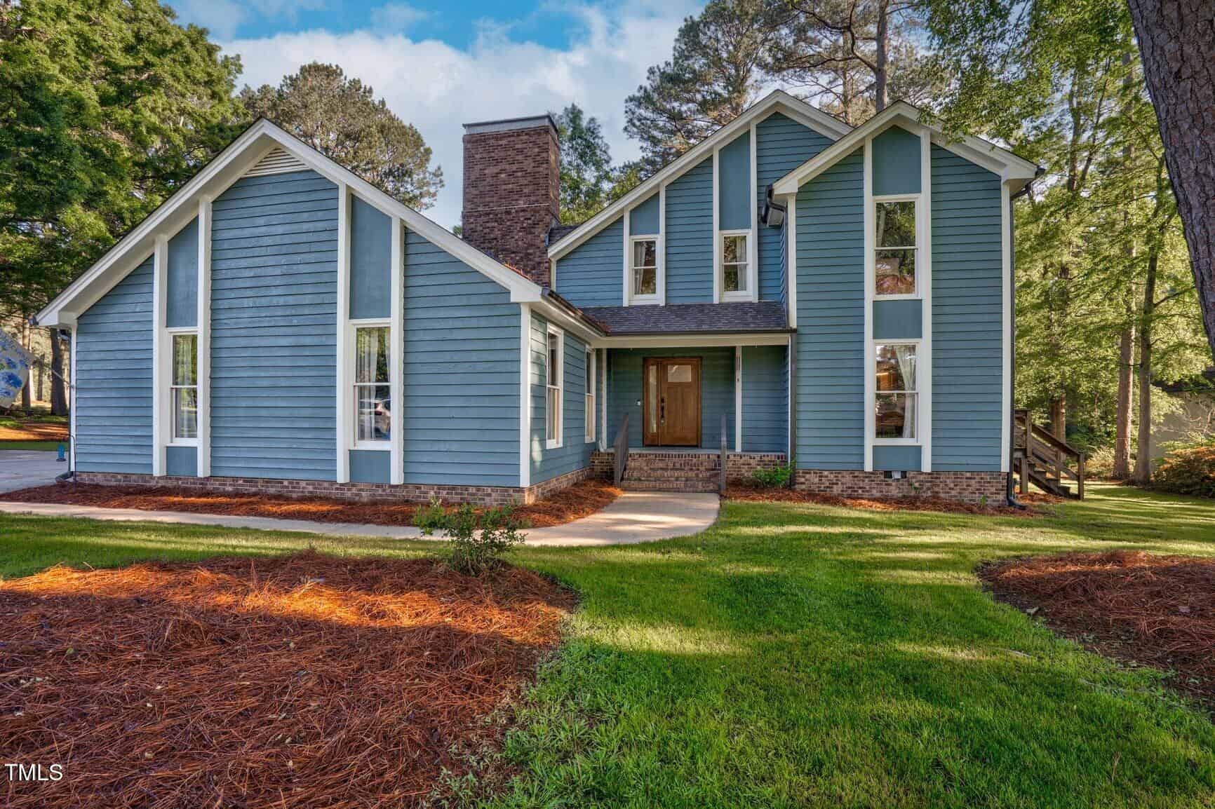 A two-story blue house with white trim, large windows, a brick chimney, and a wooden front door. The home is surrounded by green grass, pine straw mulch, and tall trees under a partly cloudy sky.
