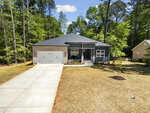 A modern single-story gray house with a two-car garage, front porch, and concrete driveway, surrounded by tall trees and a spacious yard with minimal landscaping under a blue sky.