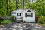 A small white house with black shutters and a front porch sits surrounded by green trees and blooming pink bushes, with a paved driveway in front.