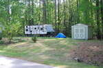 A travel trailer and a green storage shed sit on a grassy lot surrounded by tall trees. A blue tarp covers items near the shed, and the area is bordered by a road in the foreground.