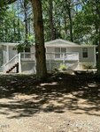 A white manufactured home with a raised front porch and white railings, surrounded by trees and shaded by sunlight, sits on a dirt lot in a wooded area.
