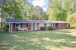 Single-story brick house with a dark roof, large front lawn, a white bench near the door, and surrounded by tall green trees under a partly cloudy sky.