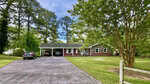 Single-story brick house with a covered carport, two cars parked, a large driveway, and surrounded by green grass, mature trees, and landscaping on a sunny day.