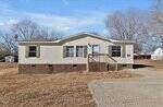 A beige, single-story manufactured home with green shutters sits on a brick foundation, featuring a small wooden porch and steps, surrounded by a dry, grassy yard under a blue sky.