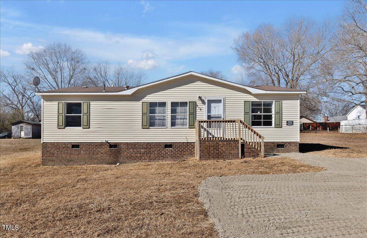 A beige, single-story manufactured home with green shutters sits on a brick foundation, featuring a small wooden porch and steps, surrounded by a dry, grassy yard under a blue sky.