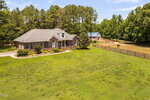 A brick house with a gray roof sits on a large grassy lot, bordered by a wooden fence. There is a barn and a horse in a fenced area, with trees and blue sky in the background.