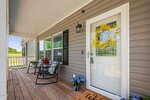A cozy front porch with wooden flooring, two black rocking chairs with cushions and flowers, a white door with a yellow wreath, blue potted hydrangeas, and a welcome mat near the entrance.