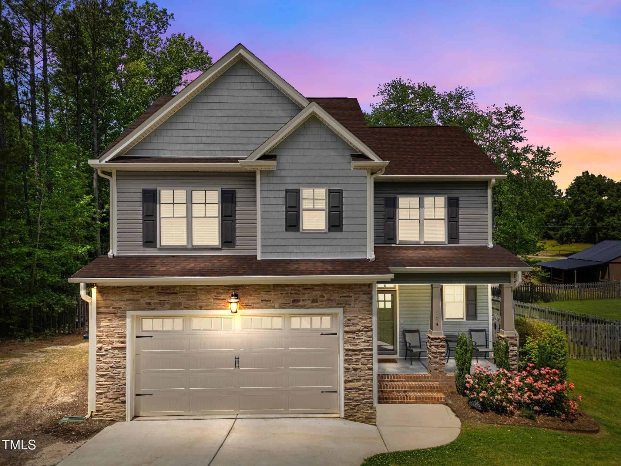 Two-story suburban house with gray siding, stone accents, and dark shutters at sunset. The driveway leads to a double garage, and a small porch with chairs is surrounded by bushes and a fenced yard. Trees are in the background.
