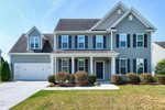 Two-story blue house with white trim, black shutters, and a covered front porch. There is a double garage on the left and neatly trimmed bushes along the front. The lawn is green, with a clear blue sky in the background.