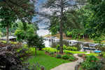 A lush green backyard with a winding stone path leads to a small dock on a calm lake, surrounded by trees and several boats moored at the shore under an overcast sky.