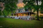 A two-story country house with a wraparound porch, white railings, and large windows sits among tall trees at sunset, with warm lights glowing inside and a grassy yard in front.