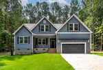 A modern two-story house with blue siding, white trim, and a stone driveway. The home features a covered front porch, double garage, and large windows, surrounded by green lawn and tall trees in the background.