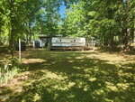 A white RV with a wooden deck is parked in a shaded, grassy yard surrounded by tall green trees. A small shed sits to the left, and sunlight filters through the leaves, casting dappled shadows on the ground.