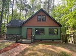 A small, two-story house with green and brown siding, a gabled roof, and a brick walkway leading to a red front door, surrounded by tall trees and greenery.