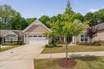 Single-story suburban house with a double garage, beige siding, and white trim. Manicured lawn, young trees, and landscaped flower beds in front. Surrounded by greenery and adjacent houses.