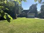 A suburban house with a dark roof, brick and siding exterior, and a two-car garage sits behind a large, sunny green lawn, surrounded by trees and neighboring homes. Leaves frame the top of the image.