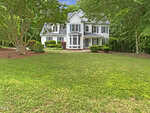 A two-story white house with black shutters sits behind a large green lawn, surrounded by trees and shrubs, under a partly cloudy sky.
