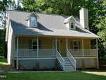 A beige two-story house with a large covered front porch, white railings, two dormer windows, a brick chimney, and surrounded by green trees under bright sunlight.