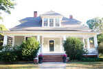 A white, two-story house with a large front porch, two brick chimneys, and a gray roof. Bushes and potted flowers line the brick steps leading to the front door. Trees surround the house, and the yard is neatly maintained.